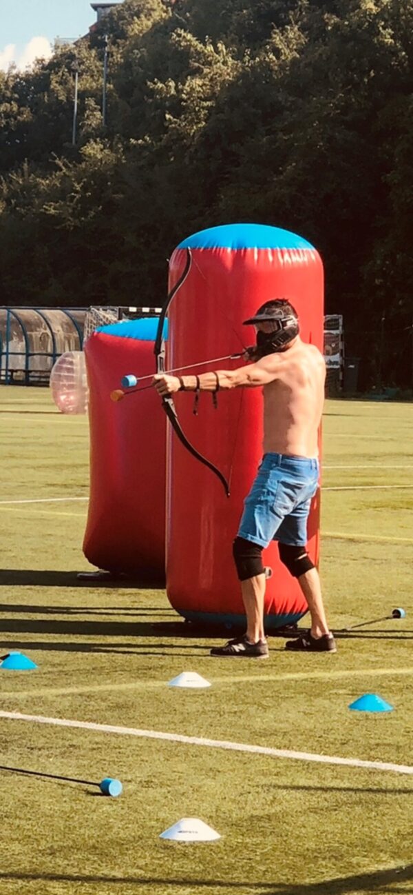 Man in shorts, and safety mask, holding a Battlezone Archery bow and arrow. taking cover behind an inflatable object.