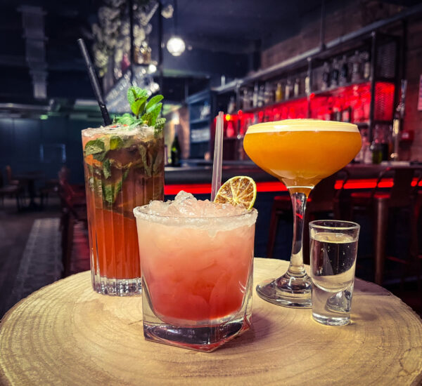 four drinks on a table, one highball collins glass with a red cocktail topped with mint leaves. one speciality cocktail glass with an orange colour topped with foam. One old fashioned glass with a pink liquid topped with ice and dried orange. one shot glass with a clear liquid. all set in Castro's newcastle for the cuban cocktail workshop.