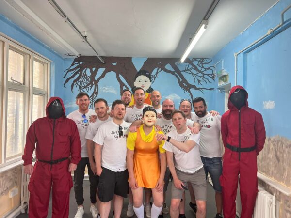 a stag party with squid game t shirts and the stag dressed as the doll. there are 2 soldiers standing to the side of the group with a mural of the doll on the wall behind.