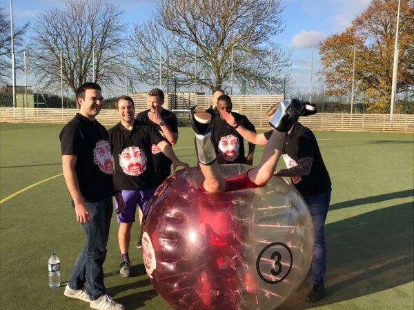 a group of men in black stag do t shirts, standing around a bubble football. Inside the bubble is a man upside down.